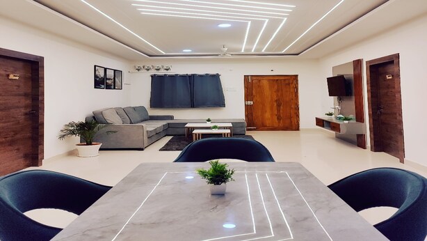 A modern living room with a sofa, a dining table with chairs, a TV, and a wooden door. - Housr Rushikonda Visakhapatnam (Visakhapatnam)