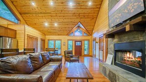 A log cabin interior with a fireplace, leather sofa, dining table, and a view of the outdoors.