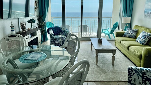 Een woonkamer met een glazen tafel, witte stoelen, een groene bank en uitzicht op de oceaan door grote ramen.