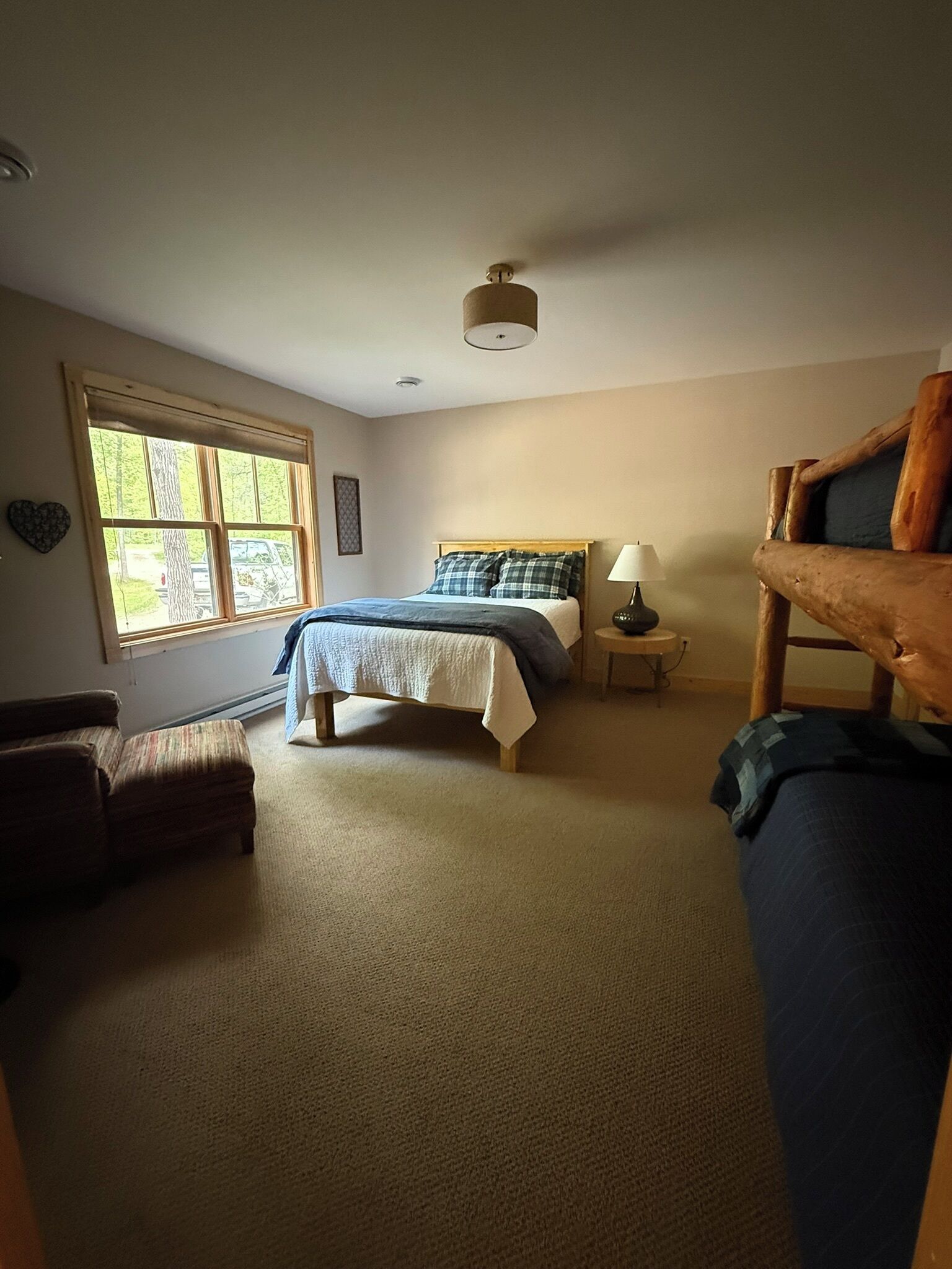 Un dormitorio con cama, mesita de noche, silla, banco de madera y ventana con vista a árboles y un vehículo.