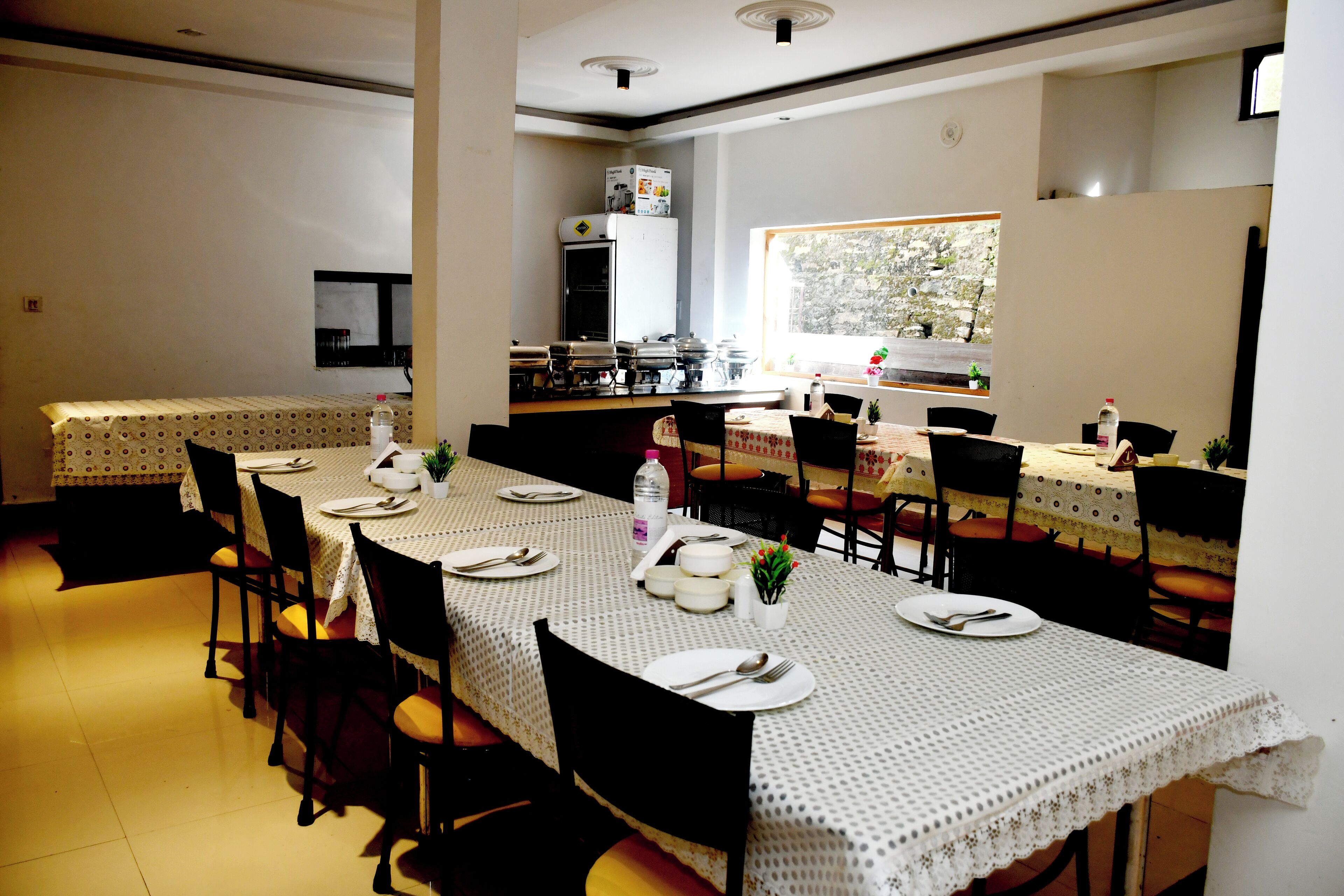A dining area with multiple tables set for meals, chairs, and a kitchen in the background.