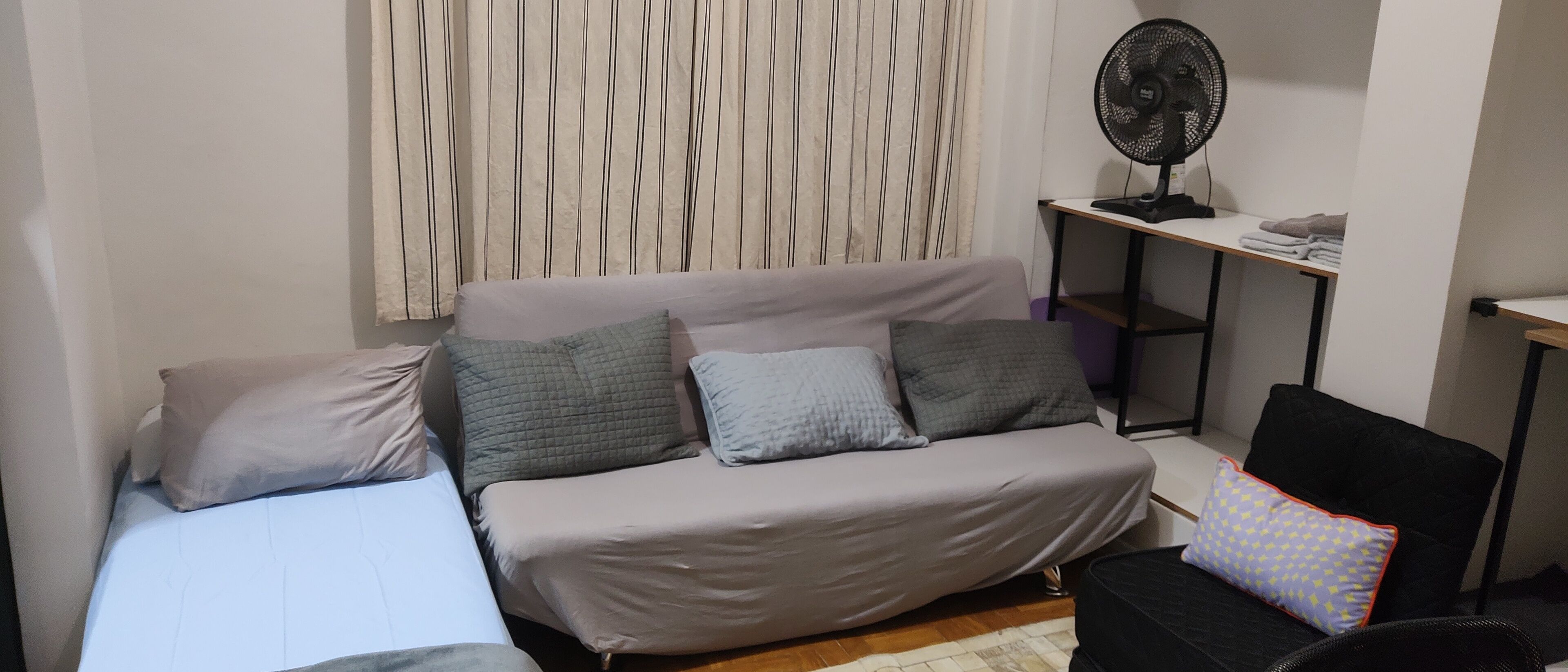 Une petite chambre à coucher, bien rangée, avec un lit, une table de chevet, une chaise et un ventilateur sur pied.