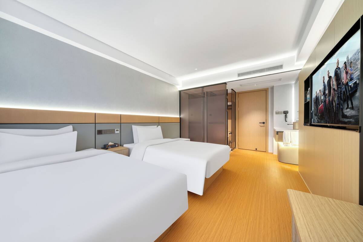 A hotel room with two beds, a wooden floor, a large painting on the wall, and a bathroom visible through a glass door.