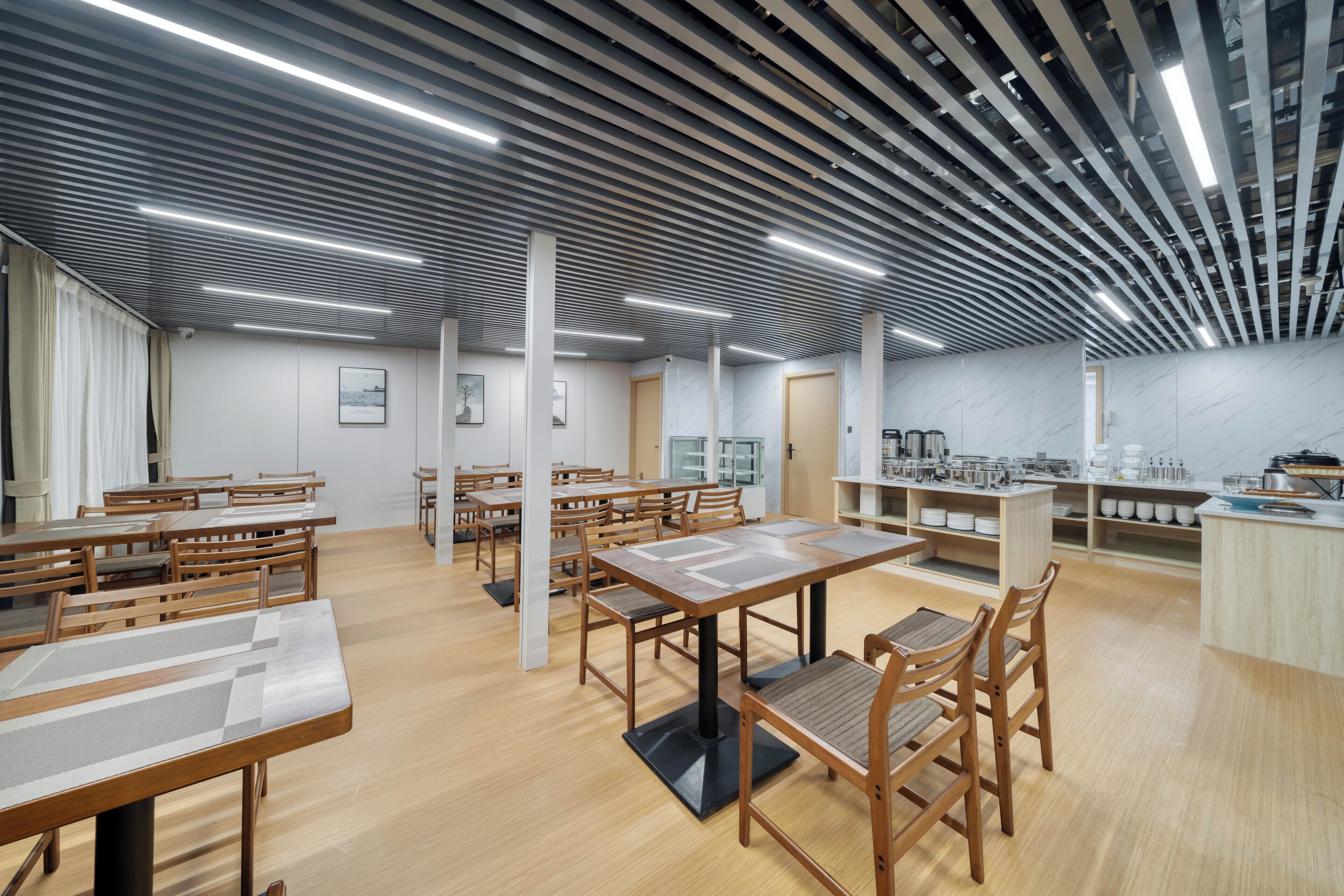 Un comedor moderno con mesas y sillas de madera, una encimera de cocina y un techo con luminarias lineales.