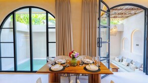 A dining area with a wooden table set for two, a vase of flowers, and a view of a pool through large windows. - Villa San Miguel by Pertama Stay (Seminyak)