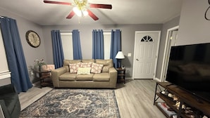 A living room with a beige sofa, a patterned rug, a ceiling fan, a TV, and a door. - Cozy Charming house in Oswego NY, with WiFi and AC (Oswego)