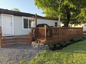 Eine Holzterrasse mit Geländer, einem Grill und einem Baum, der Schatten spendet.