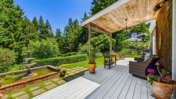 Una terraza de madera con mobiliario exterior, un jardín con una fuente y una zona cubierta con una pérgola.