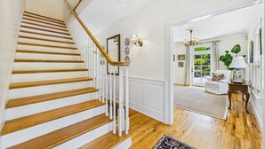 Una escalera con escalones de madera y barandillas blancas que lleva a una habitación con un ventanal grande, un sofá blanco y una araña.