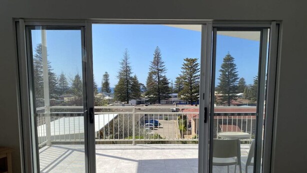 A room with a sliding glass door leading to a balcony with a view of trees and a parking lot.