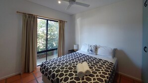 Une chambre à coucher avec un lit, un ventilateur de plafond, une fenêtre avec des rideaux et une table de chevet avec une lampe.