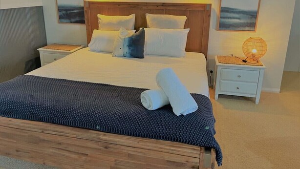 A neatly made bed with white and blue pillows, a rolled towel, and a dark blue blanket. There is a small bedside table with a lamp and a picture frame on the wall. - The Oceanaire, West Beach (West Beach)