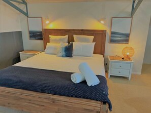 A neatly made bed with white and blue pillows, a rolled towel, and a dark blue blanket. There is a small bedside table with a lamp and a picture frame on the wall.