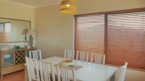 A dining area with a white table and chairs, a hanging light fixture, and a window with blinds. - The Oceanaire, West Beach (West Beach)