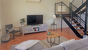 A living room with a sofa, a television, a small table, a lamp, a chair, and a staircase. - The Oceanaire, West Beach (West Beach)