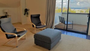 A room with a large sliding glass door leading to a balcony, two armchairs, a small ottoman, and a lamp. - The Oceanaire, West Beach (West Beach)