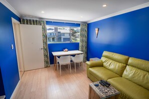 Una habitación con pared azul, un sofá amarillo, una mesita con un control remoto y una ventana con cortinas.