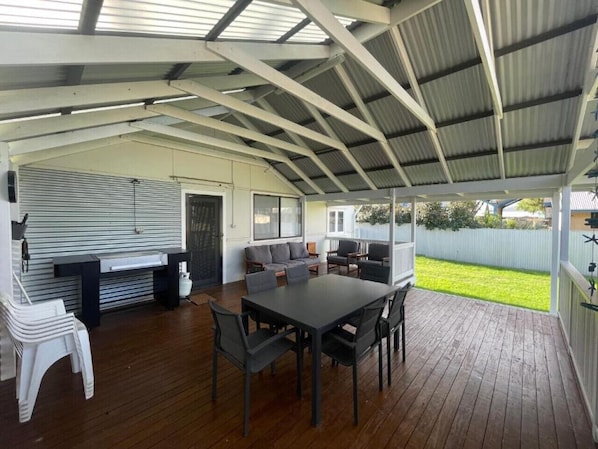 Un patio fermé avec une terrasse en bois, une table à manger avec des chaises, un canapé et un espace barbecue.