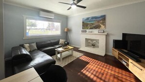 A living room with a black leather sofa, a coffee table, a fireplace, a TV stand, and a ceiling fan. - Dempster House Esperance (Esperance)