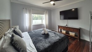 A bedroom with a bed, a wooden headboard, a bedside table, a TV mounted on the wall, and a ceiling fan. - Dempster House Esperance (Esperance)