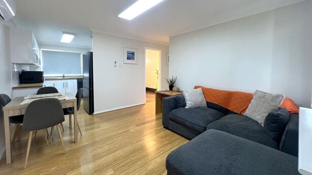 Una sala de estar moderna con un sofá gris, una mesa de comedor de madera con sillas y una cocina al fondo.