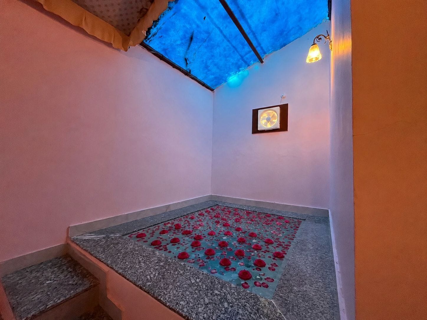 A room with a blue ceiling, a fan on the wall, and a rose petal-covered floor.