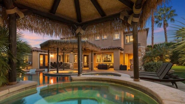Un área de piscina con techo de paja, sillas de descanso y una casa al fondo.