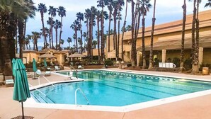Ein Swimmingpool mit Liegestühlen und Sonnenschirmen, umgeben von Palmen und Hotelgebäuden.