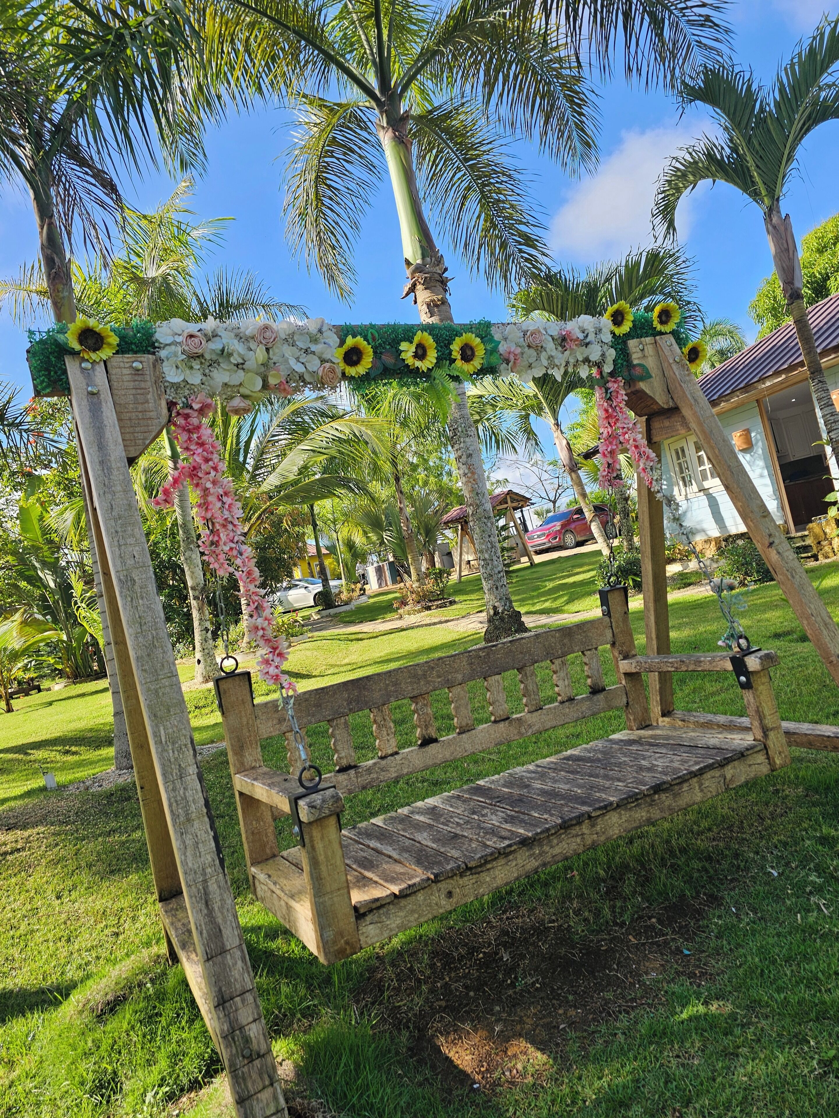 Een houten schommel met een bloemrijke boog erboven, omringd door palmbomen en een gazon.