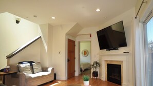 A living room with a sofa, a flat-screen TV mounted above a fireplace, and a staircase leading to another room. - Fantastic 3 bedroom, 3 bath Coastal Home Close to the Beach and Restaurants (Moss Beach)