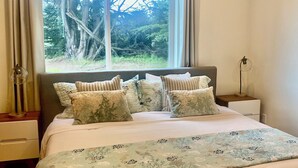 A bedroom with a large bed, multiple pillows, a nightstand, a lamp, and a window with a view of trees. - Fantastic 3 bedroom, 3 bath Coastal Home Close to the Beach and Restaurants (Moss Beach)