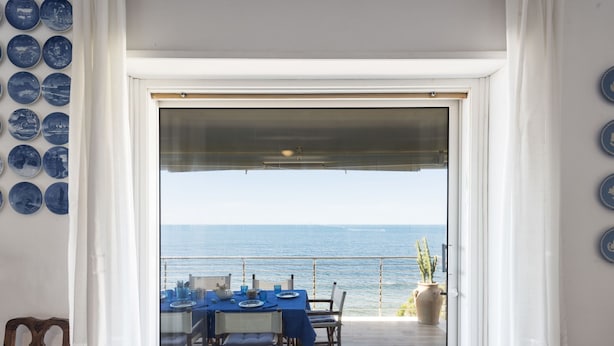 A dining area with a view of the sea, featuring a table set with blue tablecloths and chairs, a potted plant, and a wooden chair. - The Waves by Interhome (Santa Marinella)