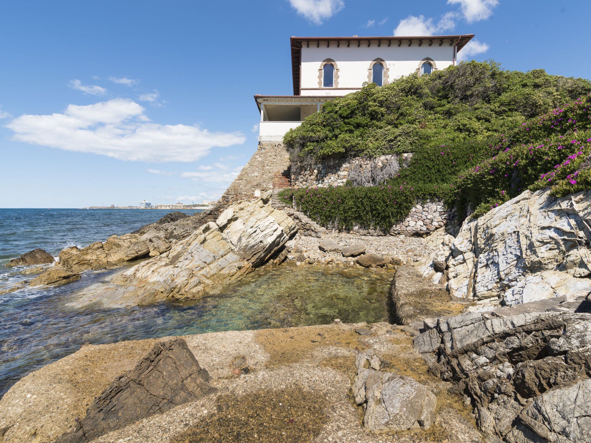 A coastal building with a white facade and arched windows, situated on a rocky cliff overlooking the sea.