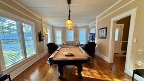 A pool table in a room with large windows, a ceiling light, and a door leading to another room. - Historic 1905 Victorian Home Near the  Rainbow River  & World Equestrian center (Dunnellon)