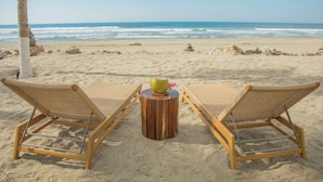 Two wicker lounge chairs on a sandy beach with a wooden stool between them, a coconut on the stool, and the ocean in the background.