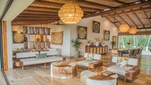 A spacious living area with a wooden ceiling, a sofa set, round ottomans, and a dining area with chairs and a table.