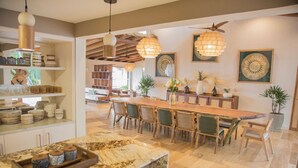 A spacious kitchen with a marble countertop, a long wooden dining table, and hanging pendant lights.