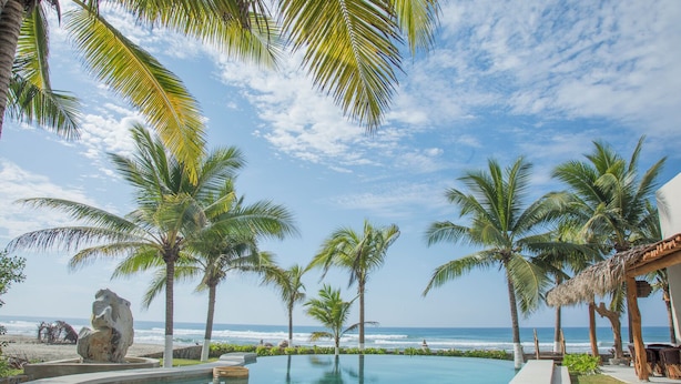 A poolside view with palm trees, a thatched structure, and a view of the ocean.