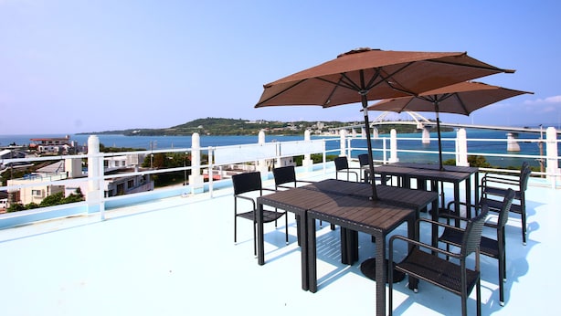 An outdoor seating area with tables and chairs, a large umbrella, and a view of the sea and a bridge. - Do pi-can - Hostel  (Motobu)
