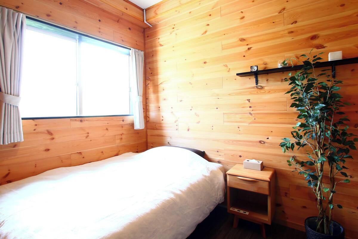 A wooden room with a bed, a nightstand, a potted plant, and a window with curtains.