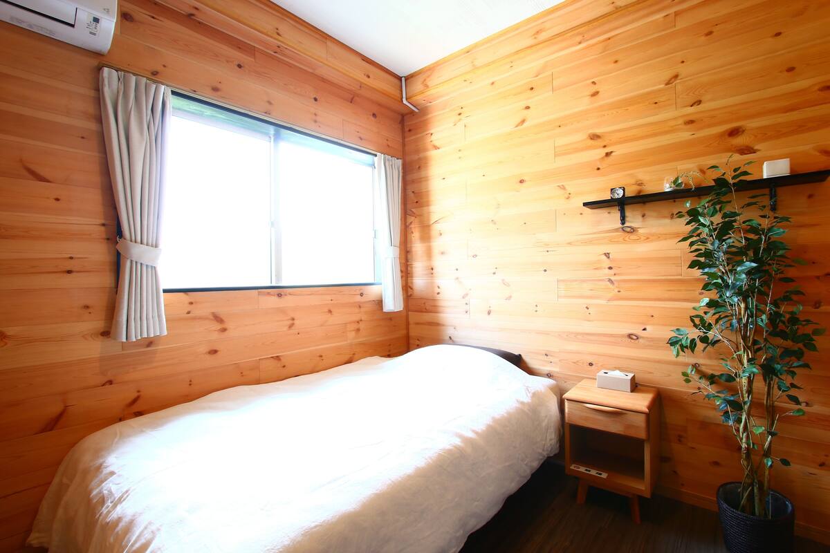 A wooden-paneled room with a bed, a window with curtains, a small bedside table, and a potted plant.