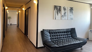 A hallway with a black leather sofa, wooden flooring, and wall-mounted artwork of palm trees. - Do pi-can - Hostel  (Motobu)