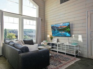 Un salon moderne avec un canapé gris, un téléviseur à écran plat fixé au mur, une petite table d’appoint ronde et un tapis décoratif.