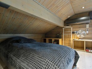 Une chambre avec un plafond en pente, un lit recouvert d’une couverture à motifs, un bureau avec un ventilateur et une étagère avec des livres et une lanterne.