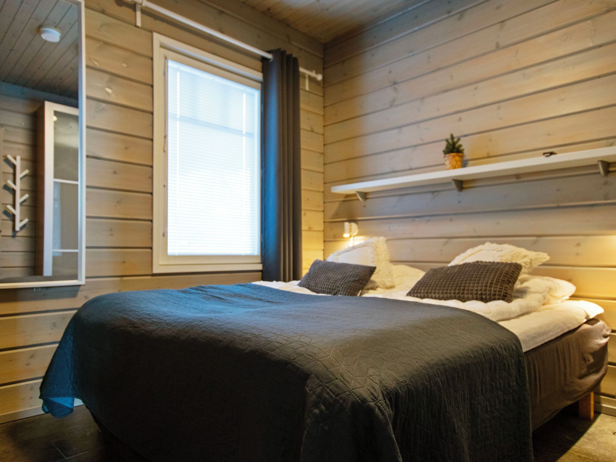 A bedroom with a wooden wall, a window, a bed with a dark bedspread, and a shelf with a plant.