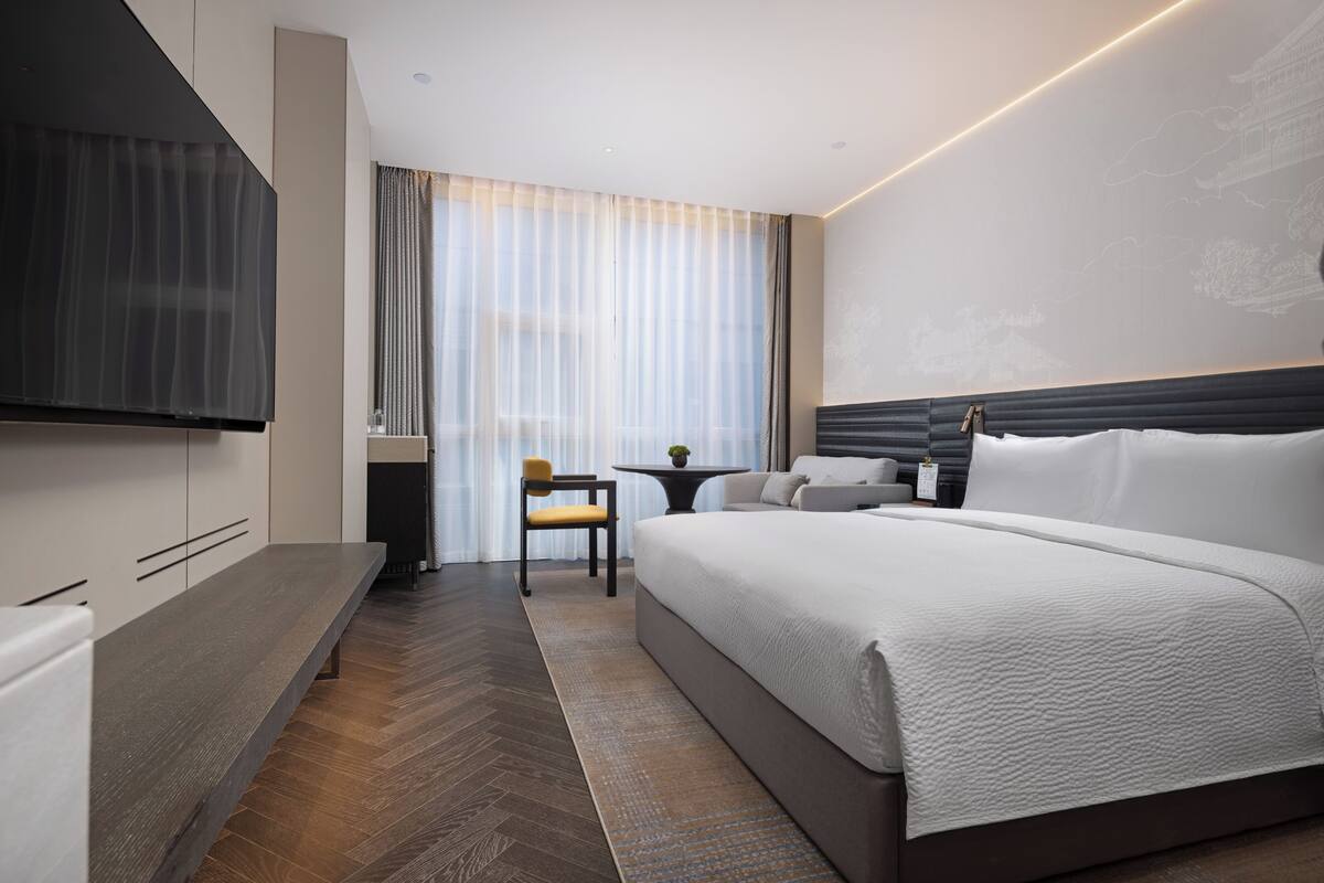 A modern hotel room with a large bed, a flat-screen TV, a desk with a chair, and a window with sheer curtains.