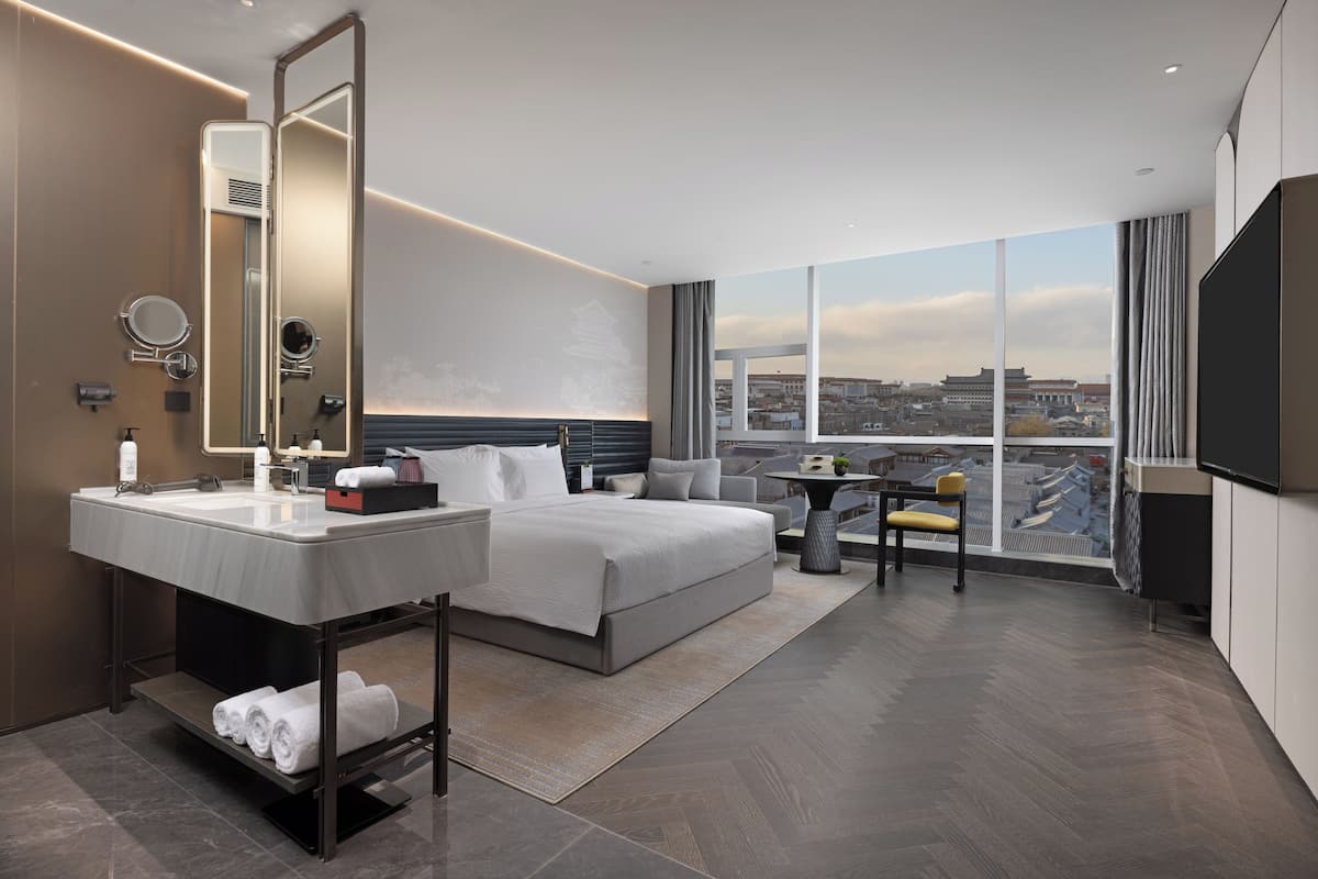 A modern hotel room with a large bed, a vanity with a mirror, a flat-screen TV, and a city view.