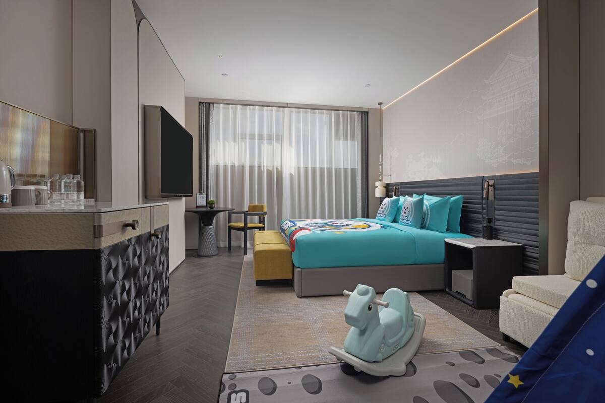 A modern hotel room with a bed, a desk, a chair, a sofa, a TV, and a decorative toy horse.
