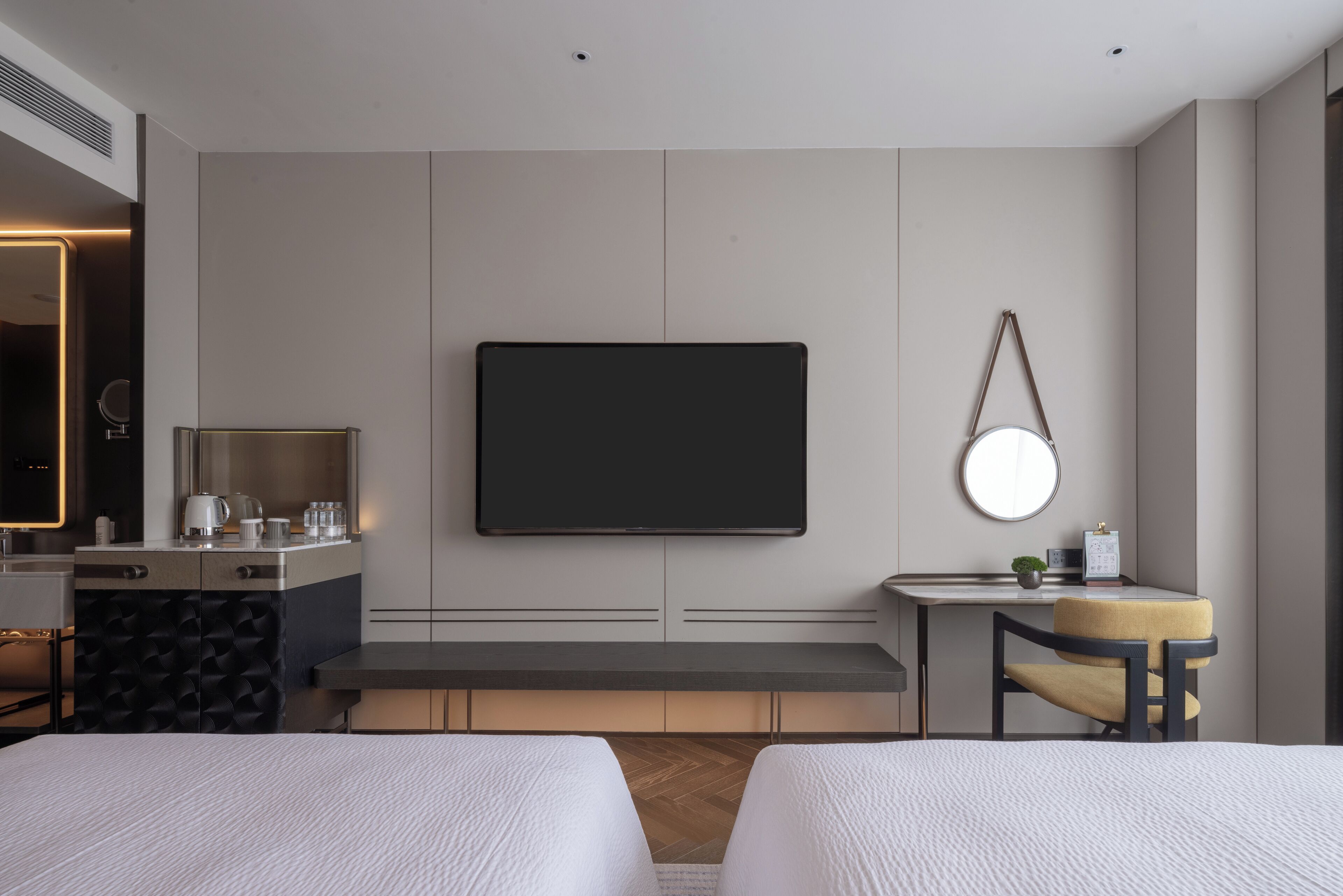 A modern hotel room with a large flat-screen TV mounted on the wall, a desk with a chair, a small round mirror, and a bed with two pillows.