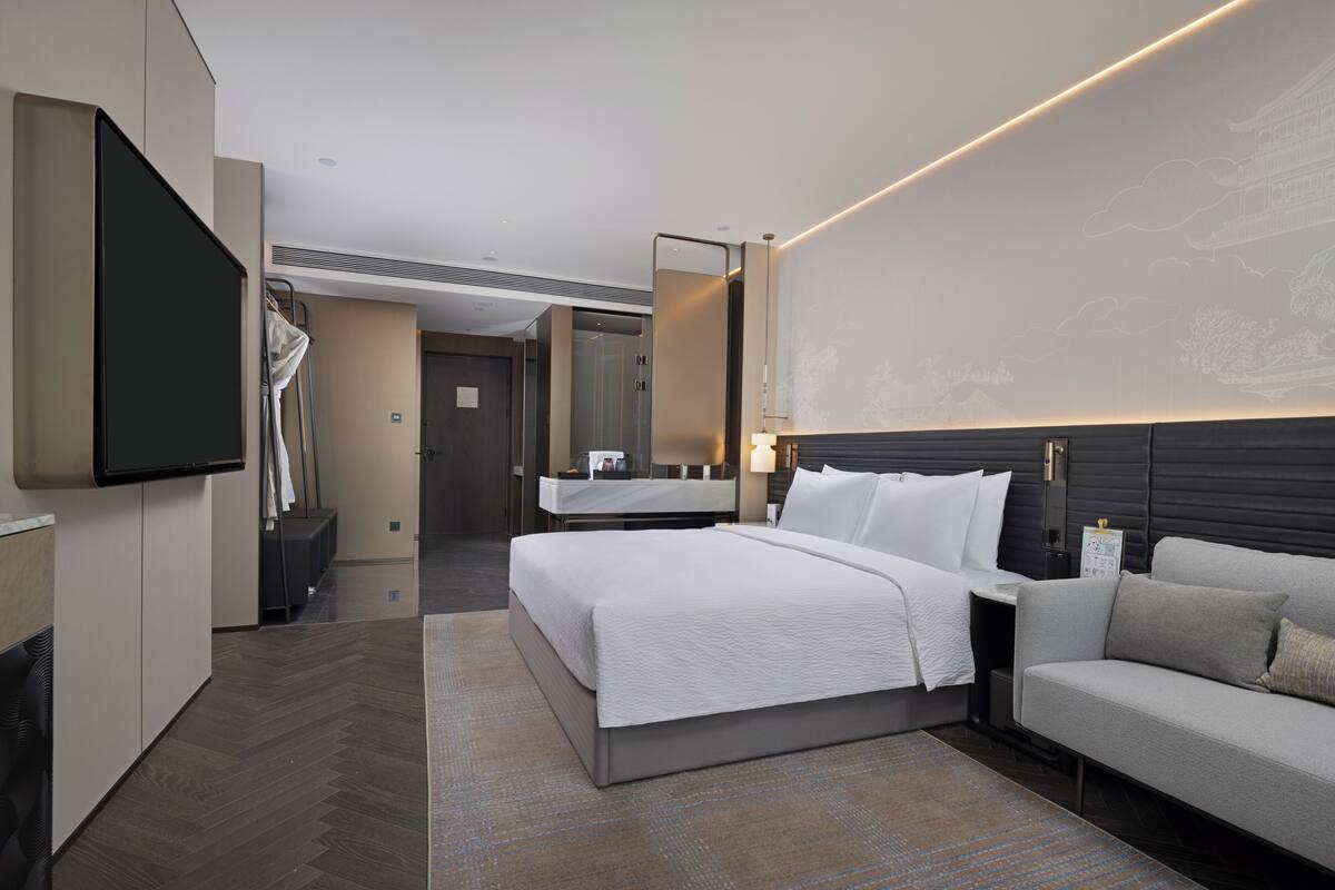 A modern hotel room with a large bed, a sofa, a flat-screen TV, a vanity with a mirror, and a wall with a subtle design.